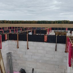 SkyDan - Budowa domu: fundamenty z bloczków, zbrojenie siatką i drewnianym szalunkiem. W tle pole i las pod pochmurnym niebem. Widoczne podpory i narzędzia.