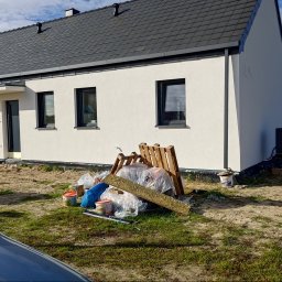 SkyDan - Nowoczesny dom jednorodzinny z białą elewacją i ciemną dachówką. Na pierwszym planie widoczne materiały budowlane i resztki po pracach wykończeniowych.