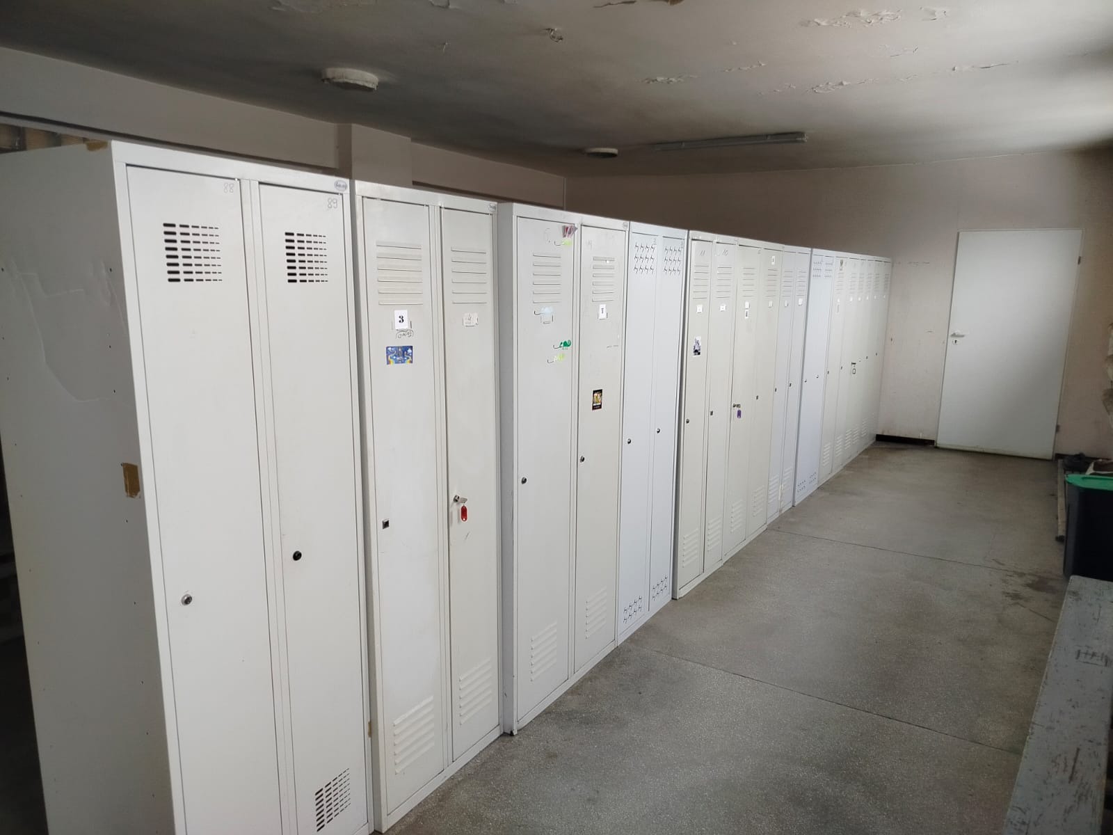 Rząd białych, metalowych szafek z numerami i naklejkami w szatni. Widoczne ślady użytkowania na szafkach i suficie. Perspektywa z dołu, oświetlenie sufitowe.