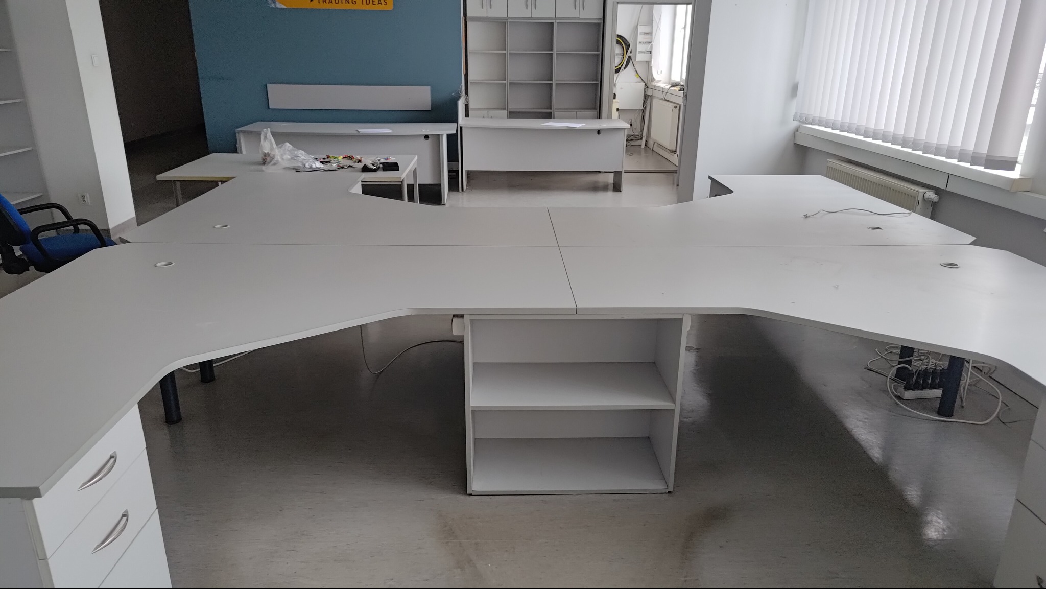 Białe biurko biurowe o nieregularnym kształcie, z szafkami i półkami, ustawione w jasnym pomieszczeniu biurowym z widocznymi instalacjami i elementami wyposażenia.