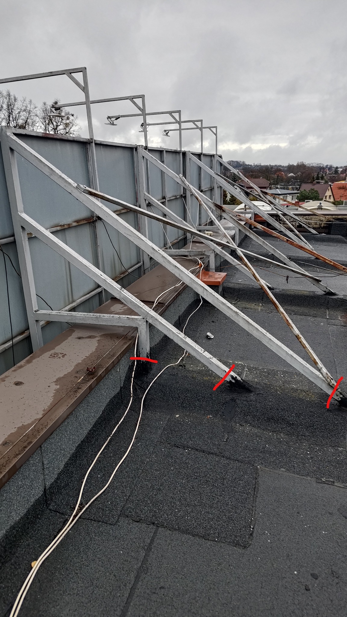 Metalowa konstrukcja wsporcza na dachu pokrytym papą, z widocznymi przewodami elektrycznymi i elementami mocującymi. W tle zabudowa miejska pod pochmurnym niebem.