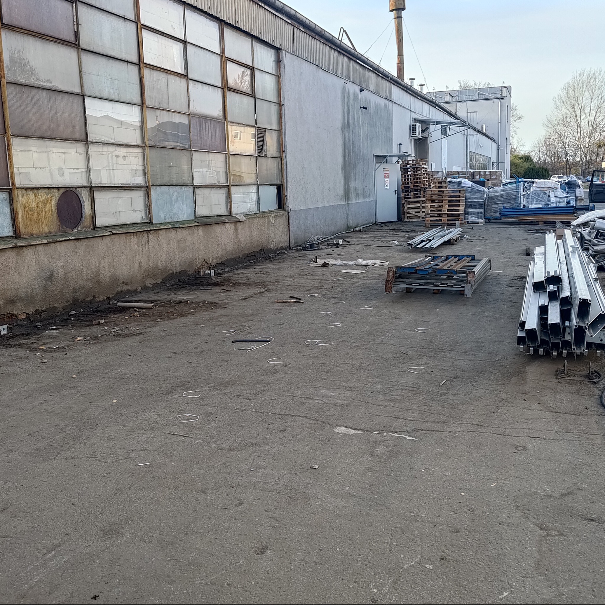 Zewnętrzny widok hali przemysłowej z rzędami okien i stertą metalowych profili na betonowym placu. Widoczne palety i elementy konstrukcyjne.
