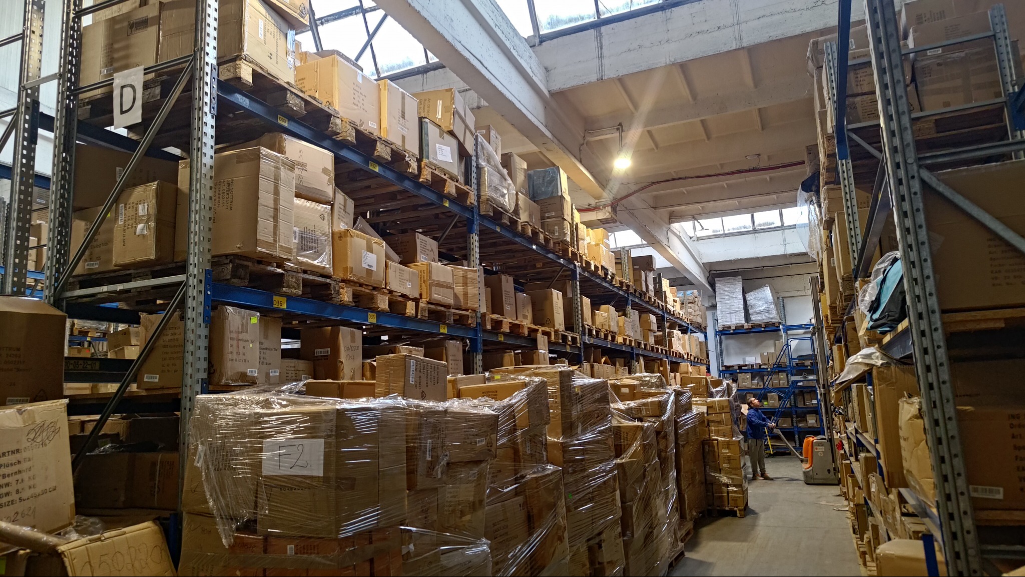 Magazyn z regałami wypełnionymi kartonami, oświetlony światłem dziennym z okien dachowych. Pracownik obsługuje wózek widłowy w wąskim korytarzu.