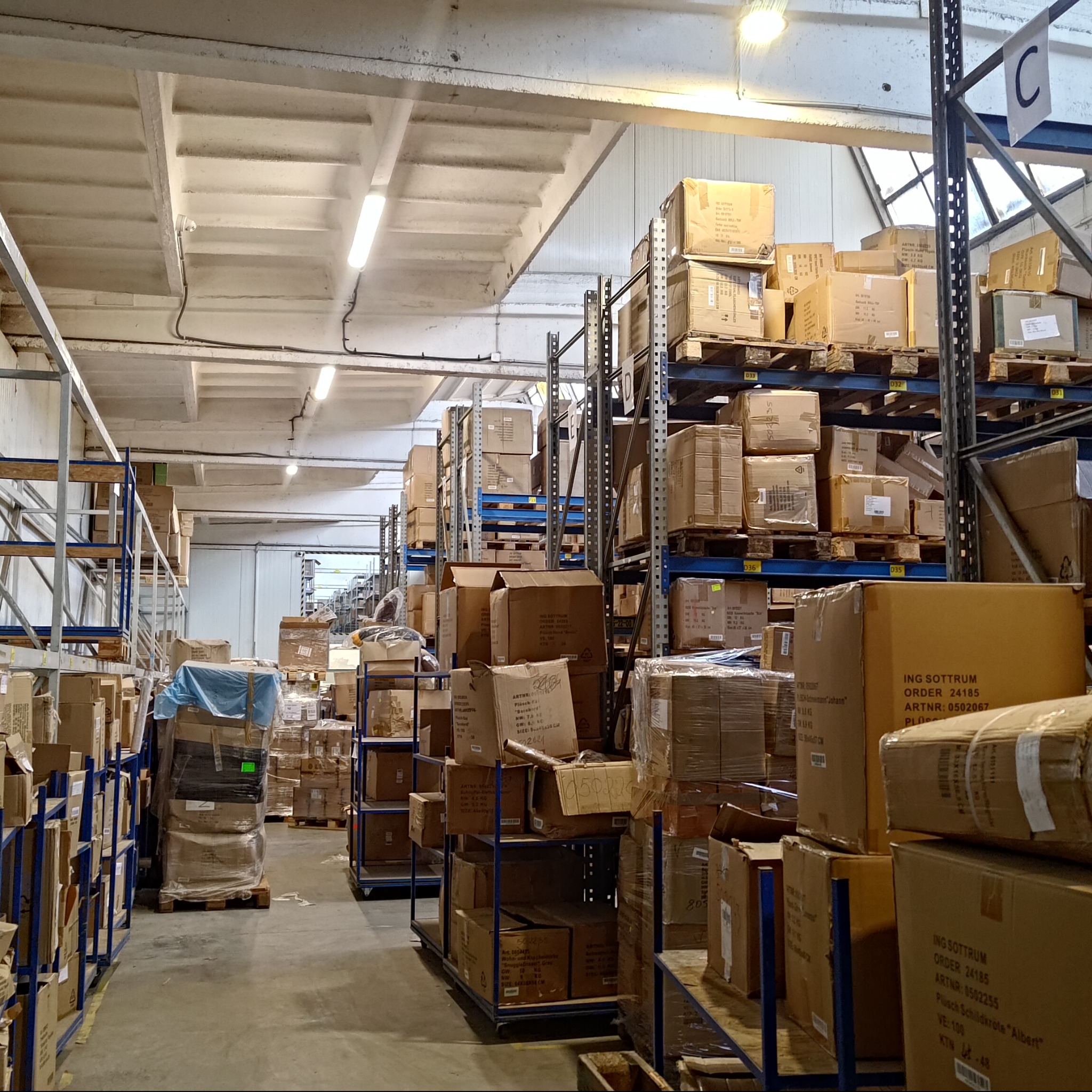 Magazyn z regałami wypełnionymi kartonami. Widoczne metalowe regały z niebieskimi elementami. Oświetlenie jarzeniowe. Perspektywa z głębi magazynu.