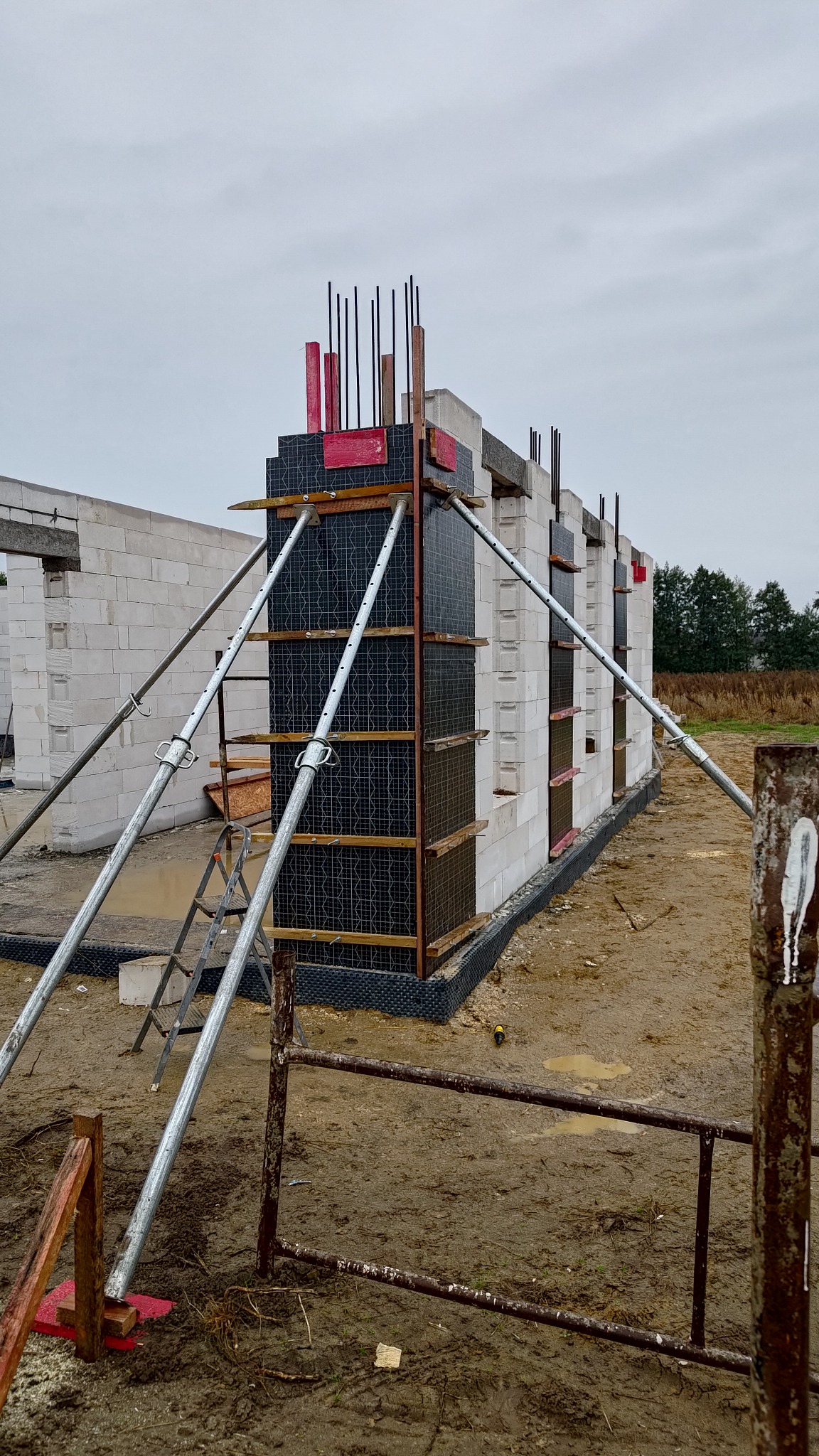 Budowa domu z betonu komórkowego: ściany w trakcie wznoszenia, wsparte metalowymi stemplami. Widoczne zbrojenia i szalunki. Tło: pochmurne niebo, teren budowy.
