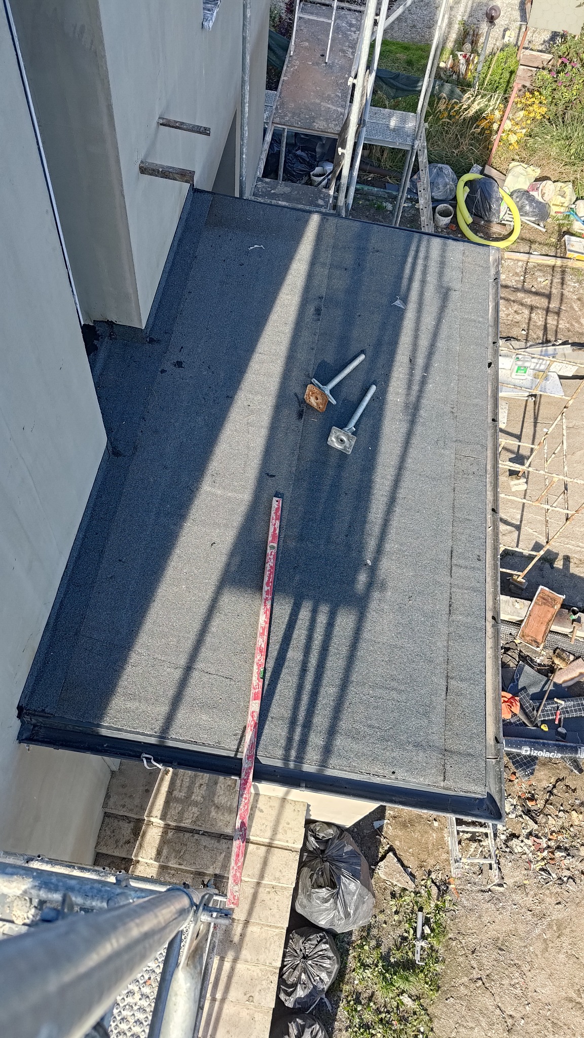 Widok z góry na płaski dach pokryty papą, z poziomnicą i elementami mocującymi. W tle rusztowanie i otoczenie budynku w słoneczny dzień.