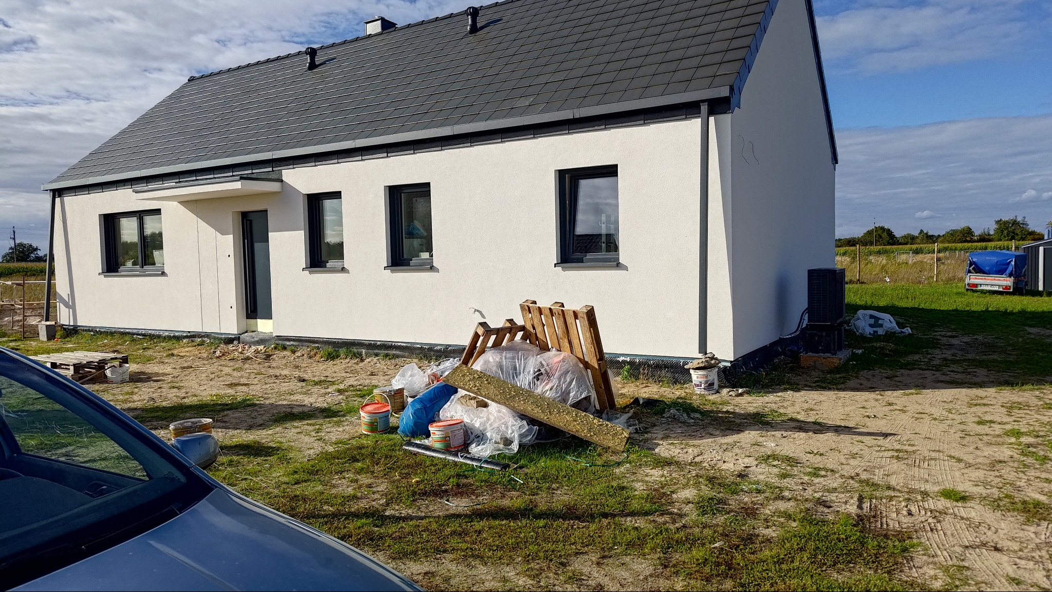 Nowoczesny dom jednorodzinny z białą elewacją i ciemną dachówką. Na pierwszym planie widoczne materiały budowlane i resztki po pracach wykończeniowych.
