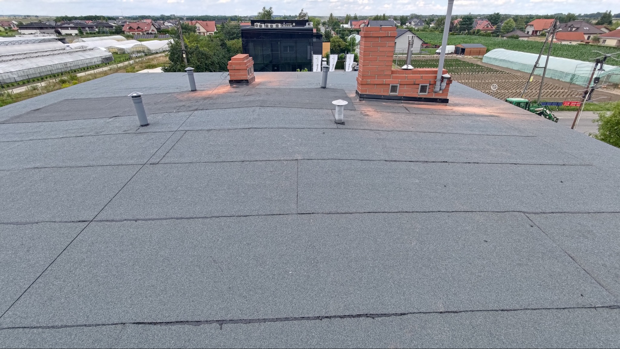 Szary, płaski dach pokryty papą termozgrzewalną z kominami z cegły i odpowietrznikami. W tle zabudowa jednorodzinna i szklarnie.