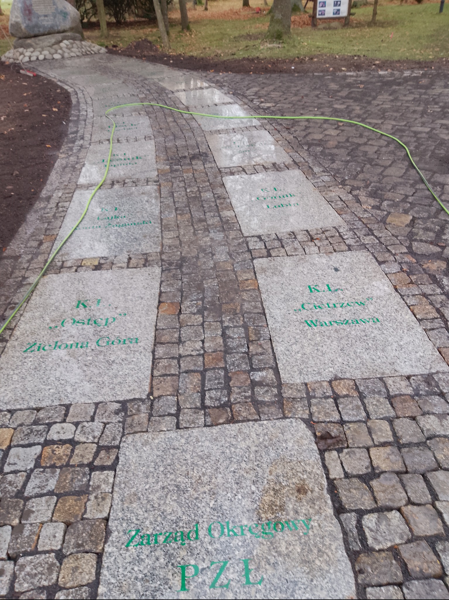 Granitowa ścieżka z wygrawerowanymi napisami w parku, otoczona kostką brukową. Widoczny fragment zielonego węża ogrodowego i drzewa w tle.