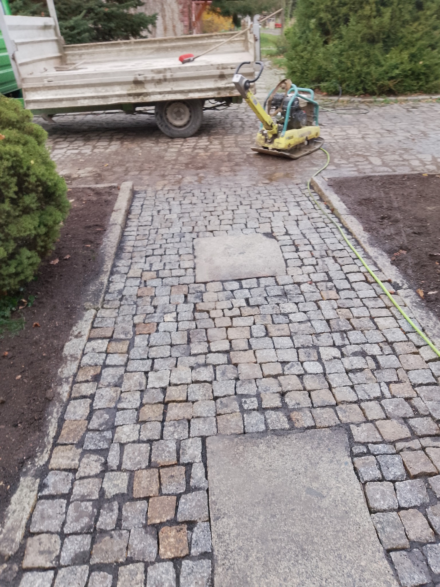 Ułożona kostka granitowa w Wymiarkach, z widoczną zagęszczarką i przyczepą. Fragment chodnika z kostki, obok rabata z ziemią i zielenią.