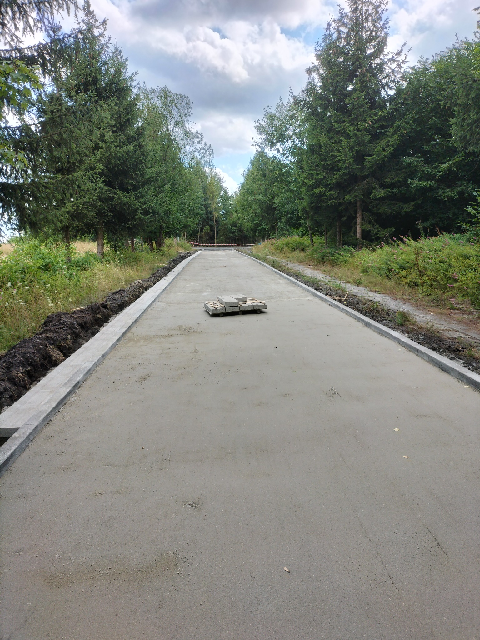 Przygotowany podkład pod kostkę brukową na drodze leśnej, z ułożonymi obrzeżami i stertą kostki w centralnej części. Widok z góry, drzewa po bokach.