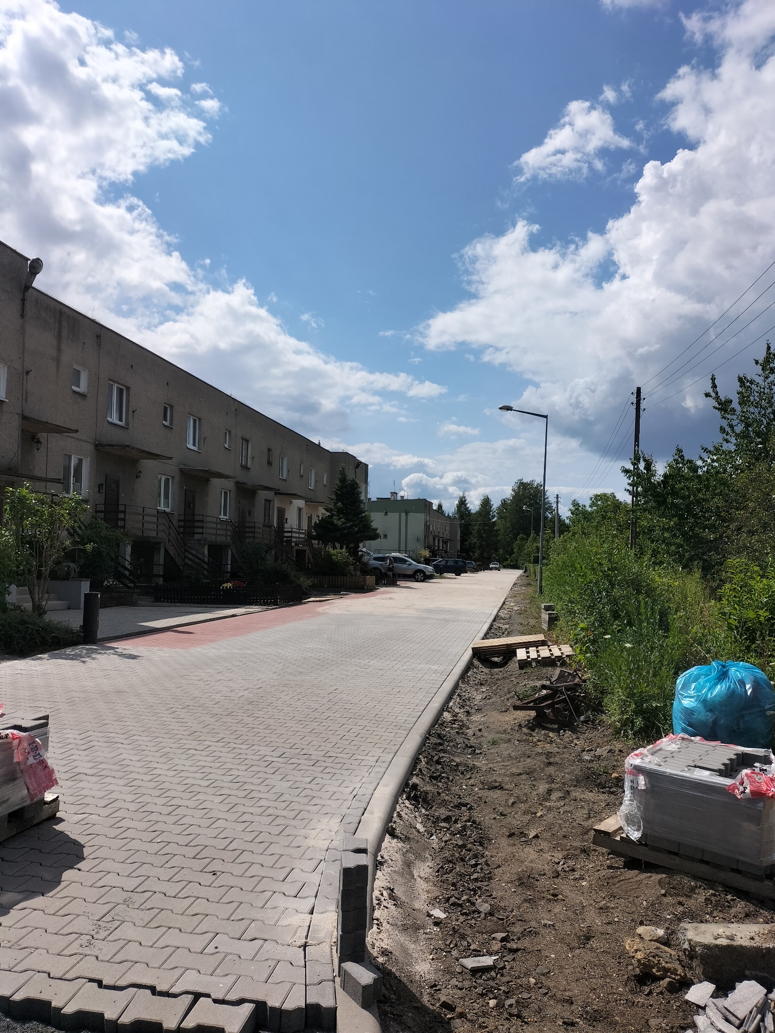 Ułożona kostka brukowa na osiedlowej drodze, widoczne palety z kostką i przygotowane podłoże. Słoneczny dzień, błękitne niebo z chmurami.