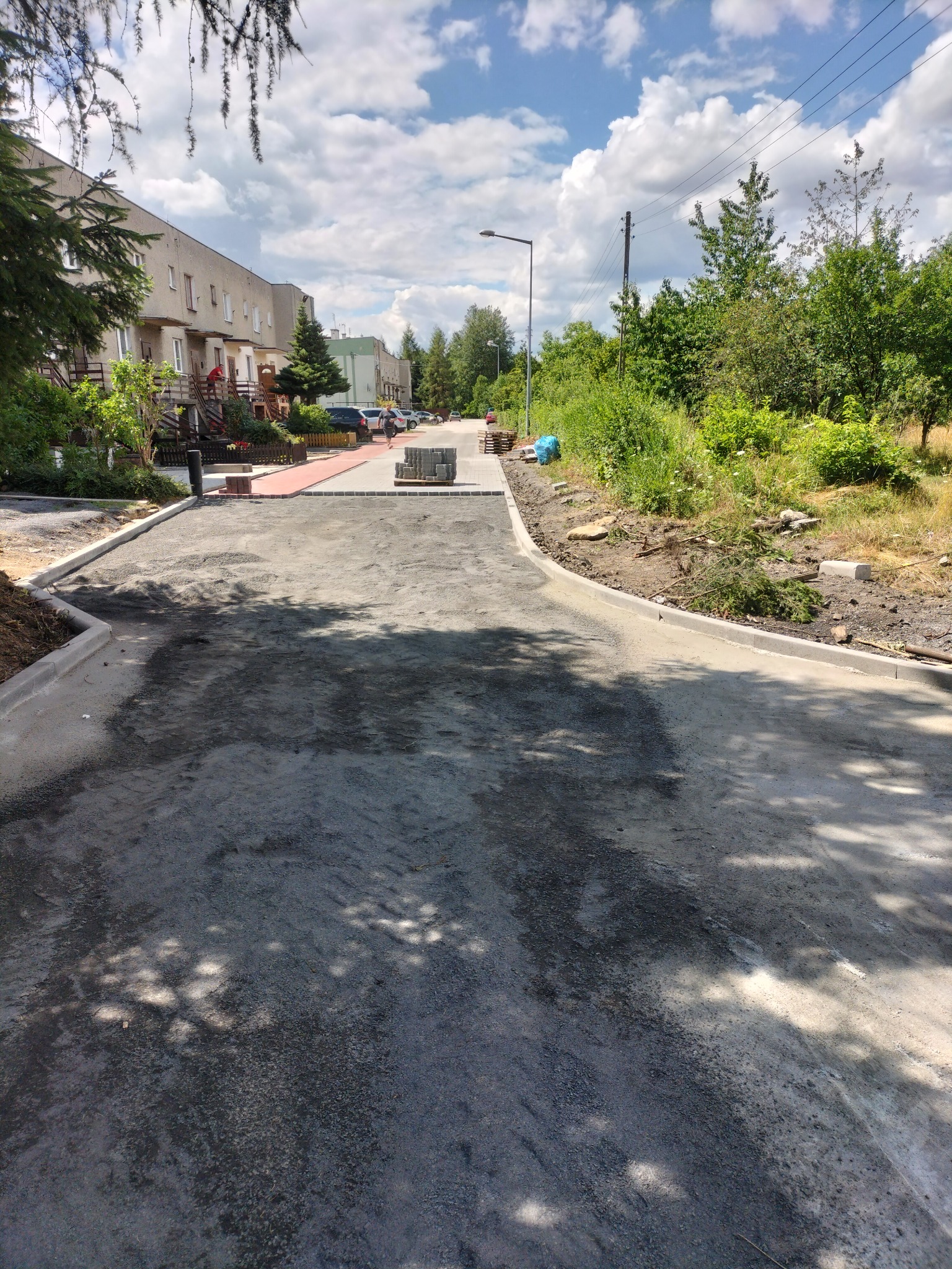 Fragment placu budowy z częściowo ułożoną czerwoną kostką brukową i przygotowanym podłożem. W tle budynki mieszkalne i błękitne niebo z chmurami.