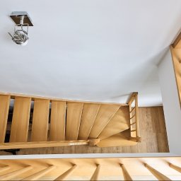 Mateusz Bojdoł - Ujęcie z góry na drewniane schody kręcone z jasnego drewna, z widocznym oświetleniem sufitowym i fragmentem podłogi. Minimalistyczny design.