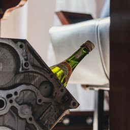 Mateusz Bojdoł - Nietypowe ujęcie butelki sake za blokiem silnika w Łaziskach Górnych. Elementy stołu i krzesła w tle. Oryginalna kompozycja z wykorzystaniem motoryzacyjnych detali.