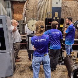 Mateusz Bojdoł - Grupa osób w koszulkach z logo firmy Polstage zwiedza zabytkową maszynownię z dużym kołem linowym i manometrami. Przewodnik objaśnia działanie urządzeń.