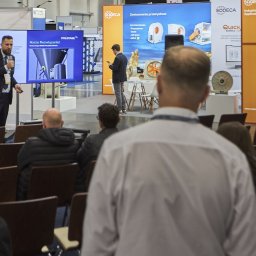 Mateusz Bojdoł - Prezentacja na targach: mężczyzna w garniturze omawia technologię dysz pulsacyjnych Polstage na ekranie. Widzowie siedzą na krzesłach, w tle stoiska firmowe.