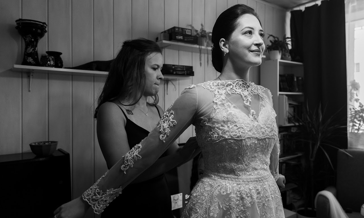 Czarno-białe zdjęcie panny młodej w koronkowej sukni, dopinanej przez kobietę w czarnej sukience, w pokoju z półkami i dekoracjami, w trakcie przygotowań ślubnych.