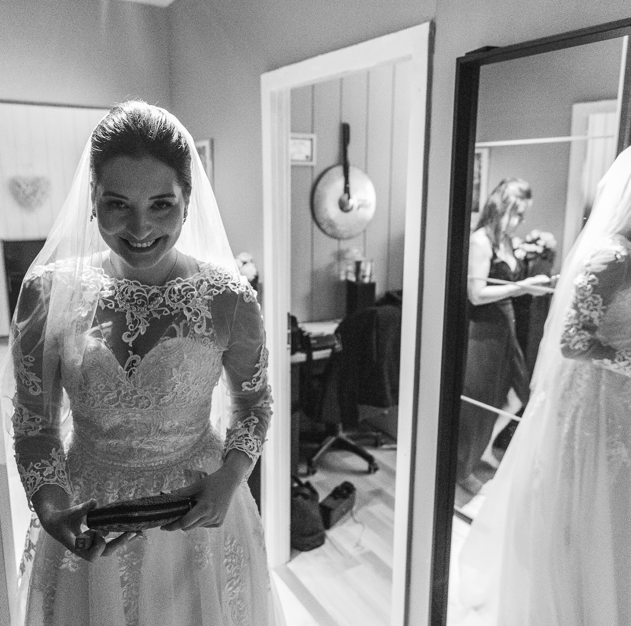 Uśmiechnięta panna młoda w koronkowej sukni i welonie, trzymająca małą torebkę. W tle lustro odbijające kobietę z bukietem i fragment sukni ślubnej.