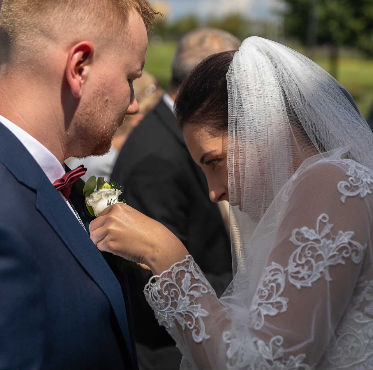 Panna młoda przypina butonierkę do granatowego garnituru pana młodego. Delikatna koronka na welonie i rękawach sukni. Ujęcie z bliska, w tle zieleń.