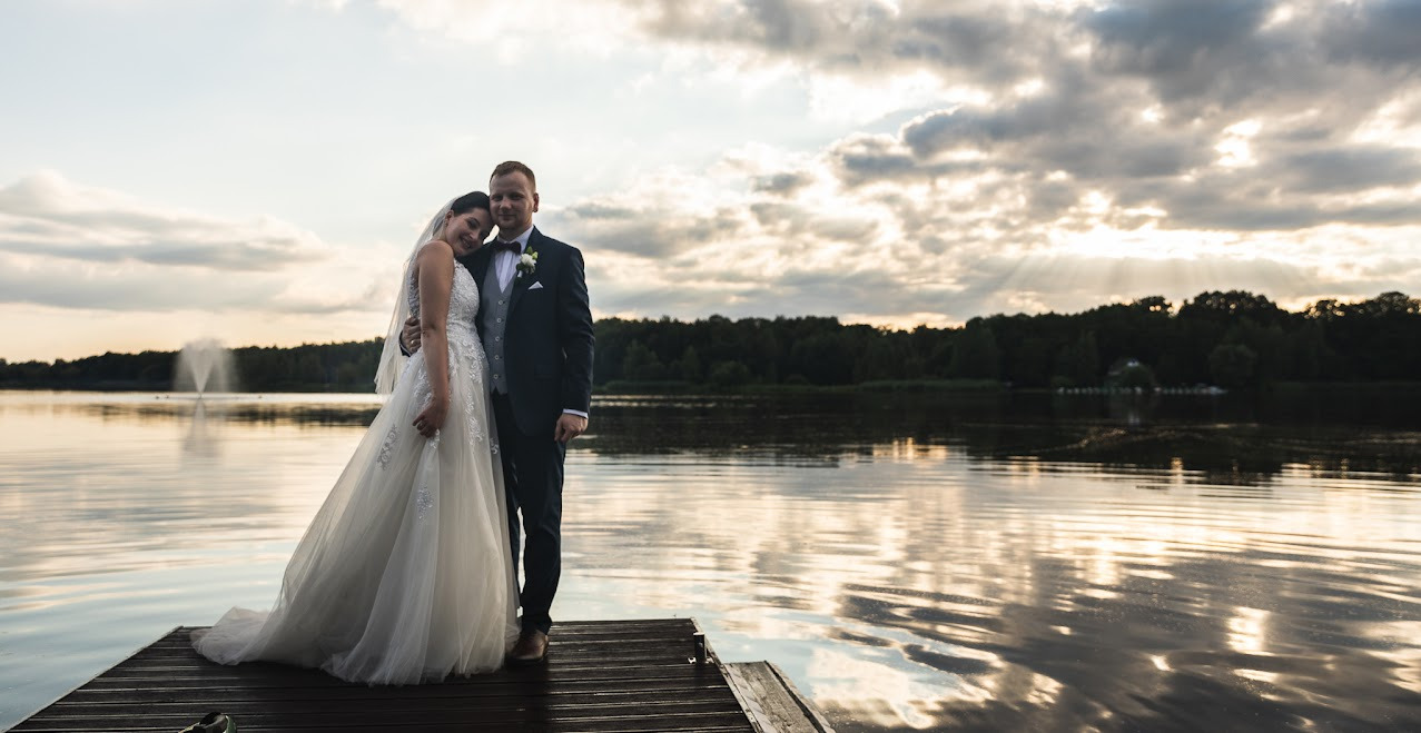 Para młoda w eleganckich strojach pozuje na drewnianym pomoście nad jeziorem o zachodzie słońca. W tle fontanna i las, a na niebie promienie przebijające się przez chmury.