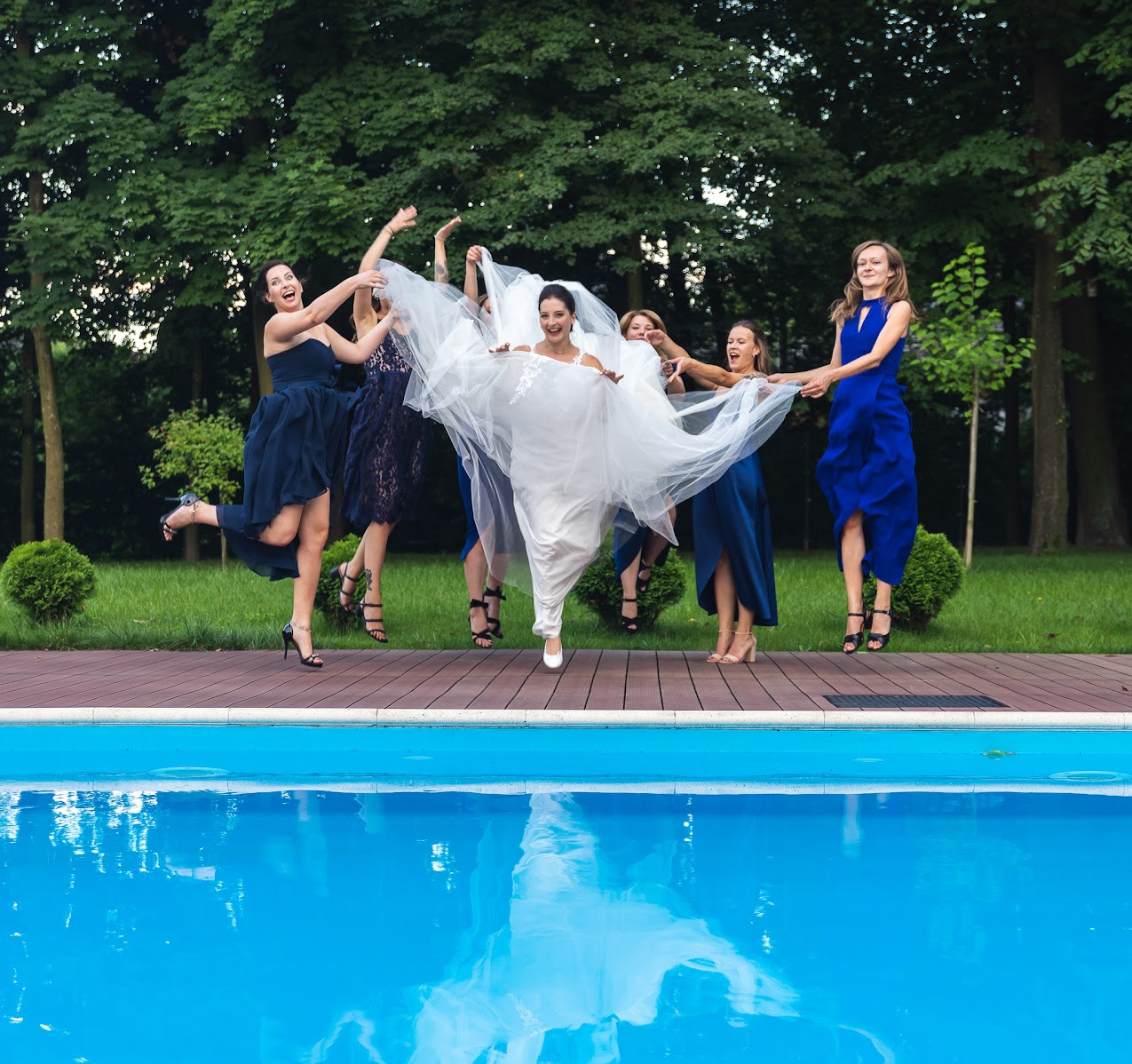 Radosna panna młoda w białej sukni, otoczona druhnami w niebieskich sukienkach, skacze przy basenie. W tle zielony trawnik i drzewa. Dynamiczne ujęcie.