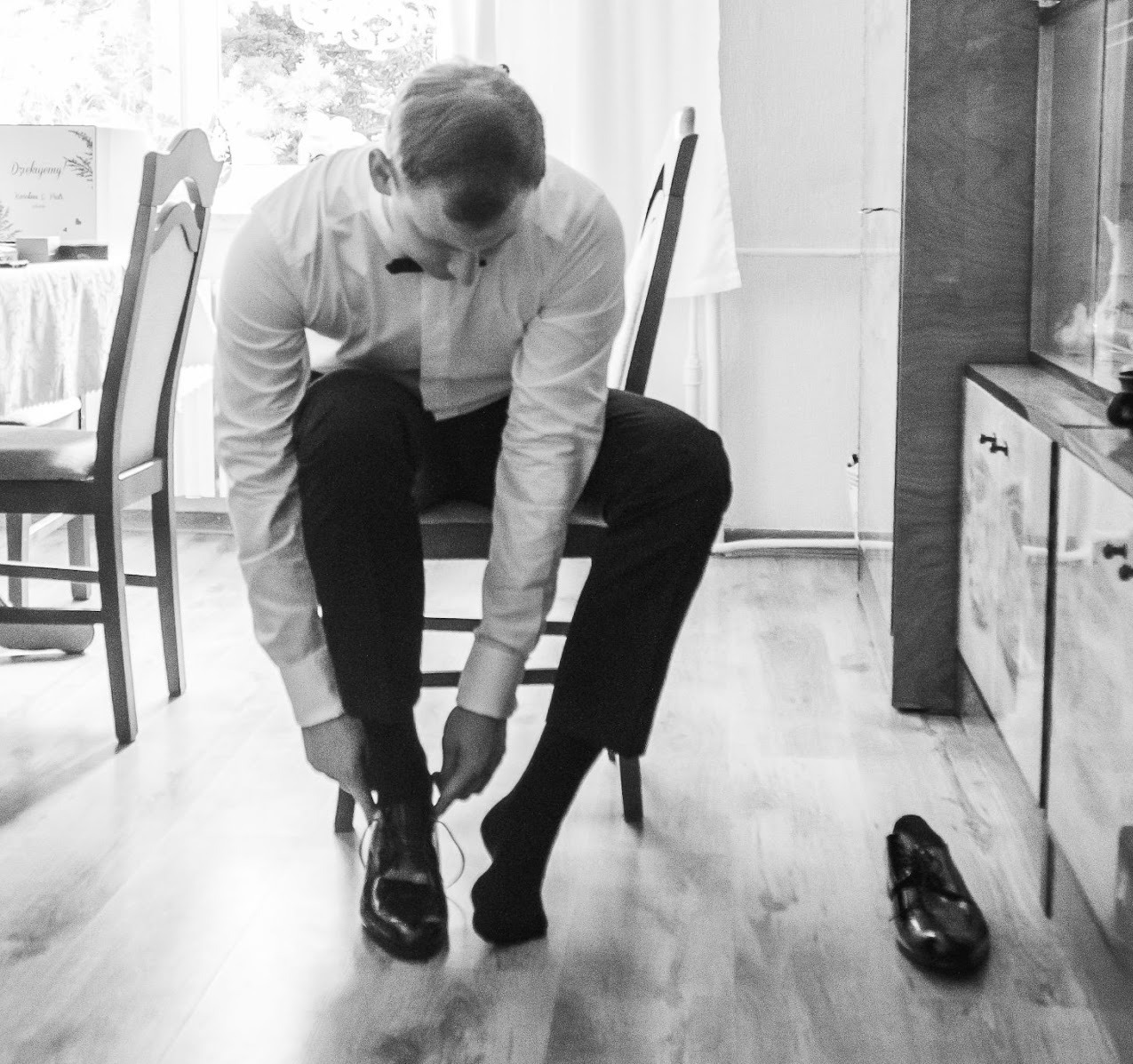 Czarno-białe zdjęcie mężczyzny w koszuli i muszce, siedzącego na krześle i wiążącego sznurowadła butów. Obok leży drugi but. W tle stół z dekoracją.