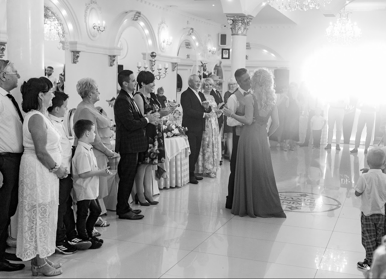 Czarno-białe zdjęcie z sali balowej: para młoda tańczy, otoczona gośćmi. Dekoracje, kolumny i parkiet dodają elegancji. Jasne światło tworzy subtelny efekt.