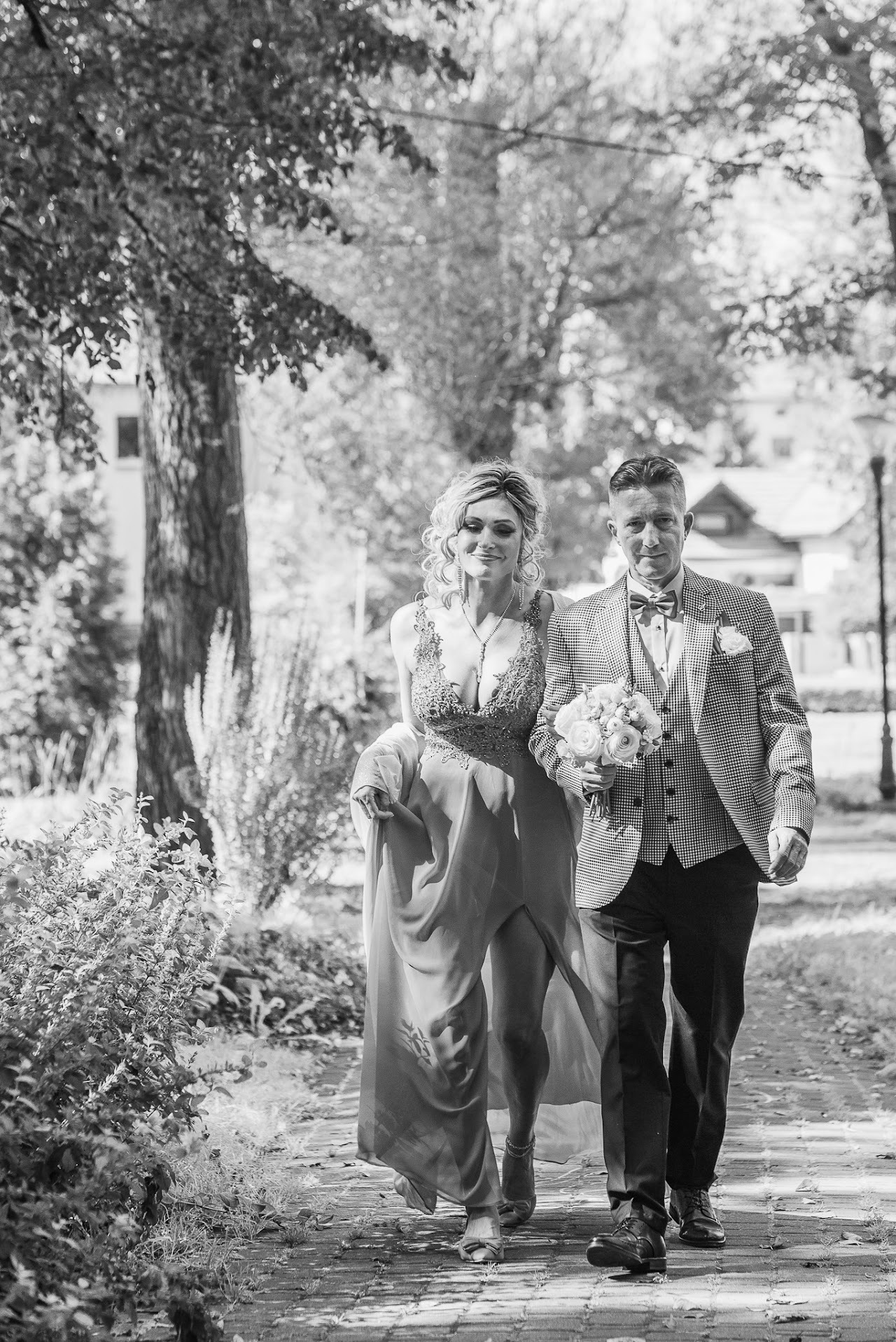 Czarno-białe zdjęcie pary młodej spacerującej po parku. Panna młoda w sukni z koronką, pan młody w marynarce w kratę i muszce. Bukiet w ręku. Romantyczna atmosfera.