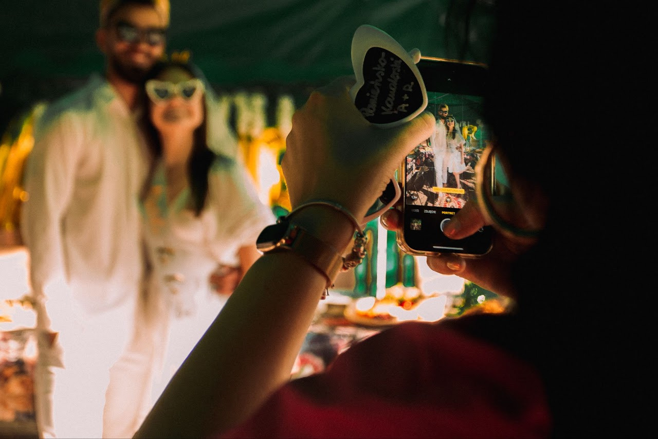 Gość weselny robi telefonem zdjęcie młodej parze w białych strojach, trzymając tabliczkę z imionami nowożeńców. Ujęcie zza pleców fotografa.