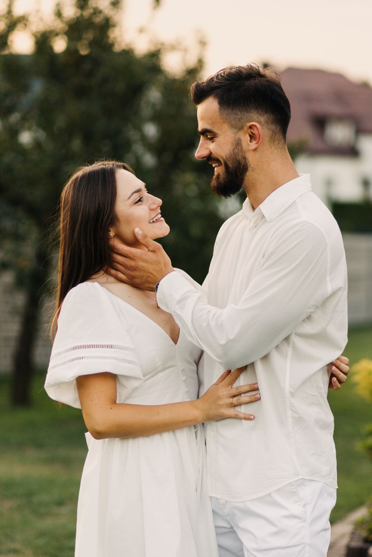 Uśmiechnięta para w bieli, on obejmuje ją, dotykając szyi, ona trzyma jego rękę. Romantyczna sesja w plenerze, naturalne światło, w tle zieleń i budynek.