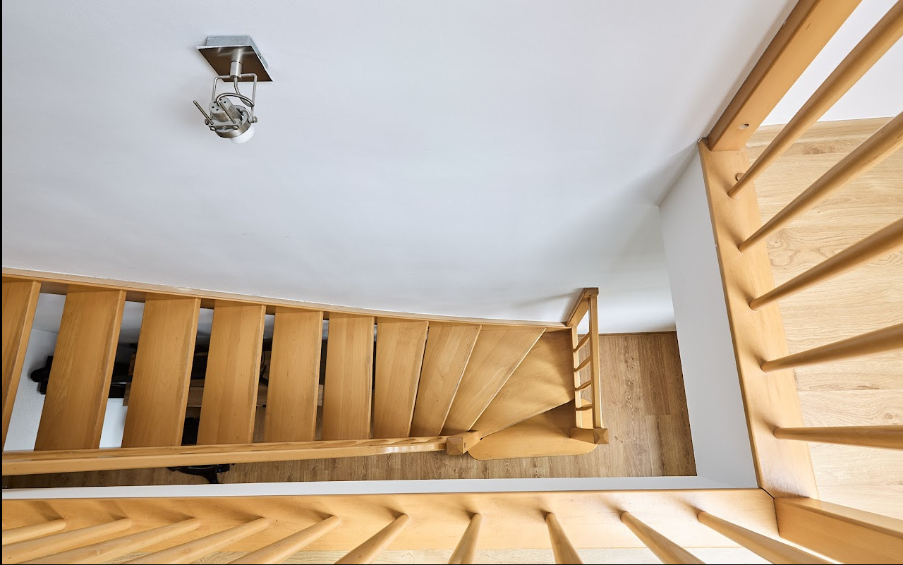 Ujęcie z góry na drewniane schody kręcone z jasnego drewna, z widocznym oświetleniem sufitowym i fragmentem podłogi. Minimalistyczny design.