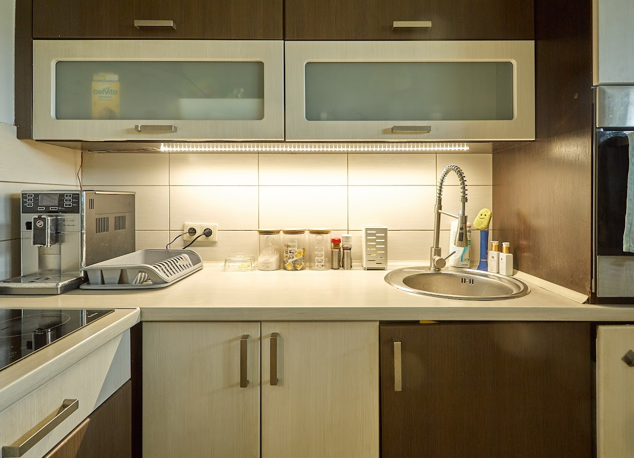 Jasna kuchnia z drewnianymi szafkami, oświetleniem LED pod szafkami i srebrnym zlewem. Na blacie widoczne akcesoria kuchenne i ekspres do kawy.