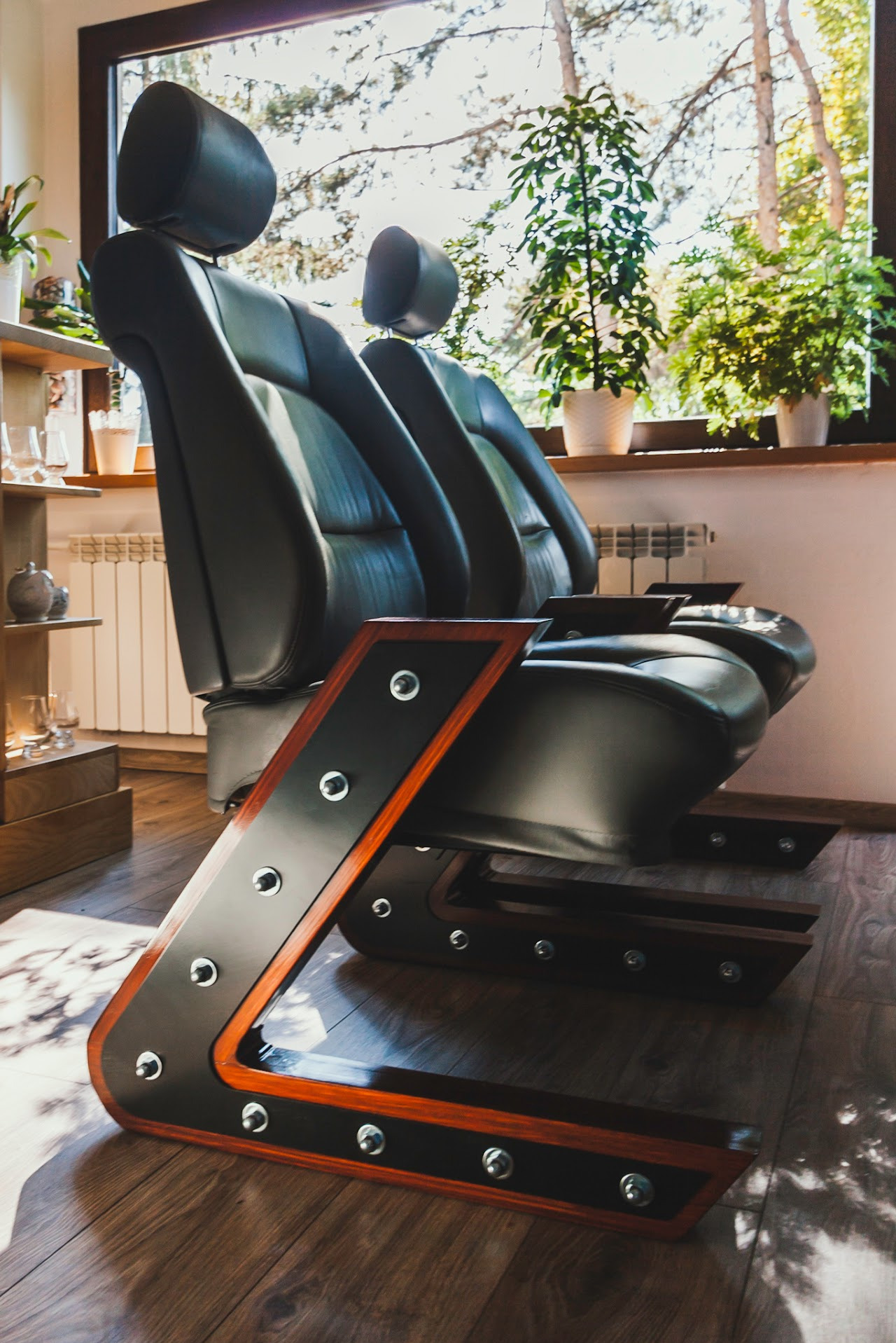 Dwa czarne, skórzane fotele z drewnianą podstawą w kształcie litery Z, zdobione śrubami, stoją na drewnianej podłodze, a w tle widać okno z roślinami.