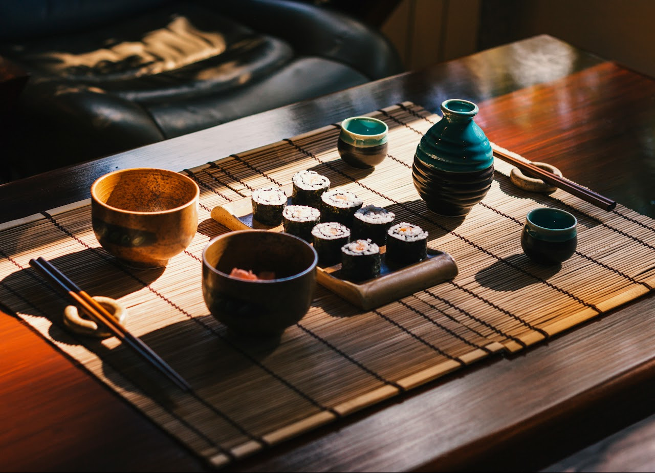 Zestaw sushi z naczyniami na bambusowej macie, oświetlony ciepłym światłem. Widoczne rolki sushi, miseczki, dzbanek na sake i pałeczki. Ujęcie z góry, podkreślające detale.
