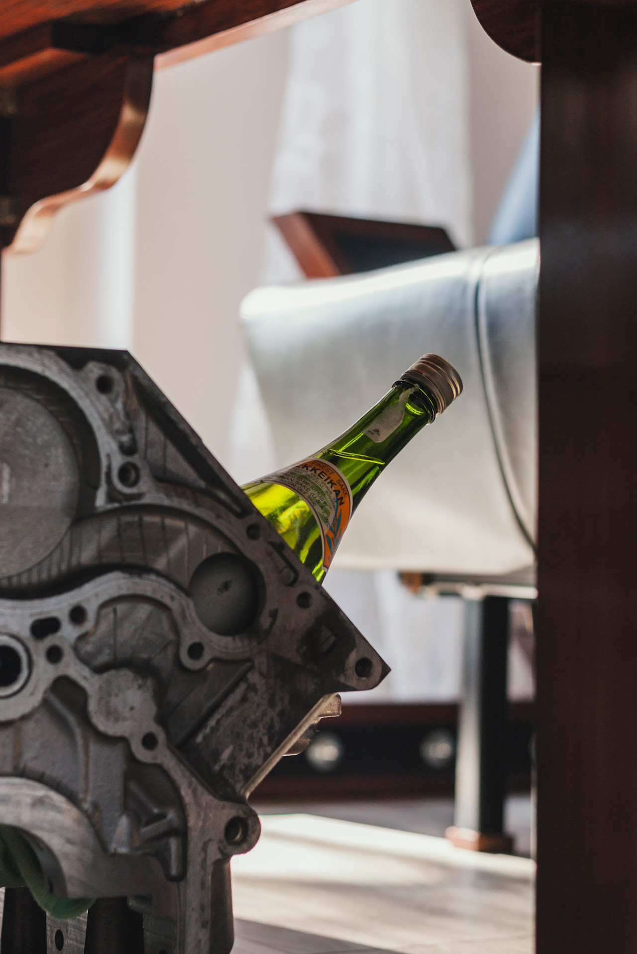 Nietypowe ujęcie butelki sake za blokiem silnika w Łaziskach Górnych. Elementy stołu i krzesła w tle. Oryginalna kompozycja z wykorzystaniem motoryzacyjnych detali.