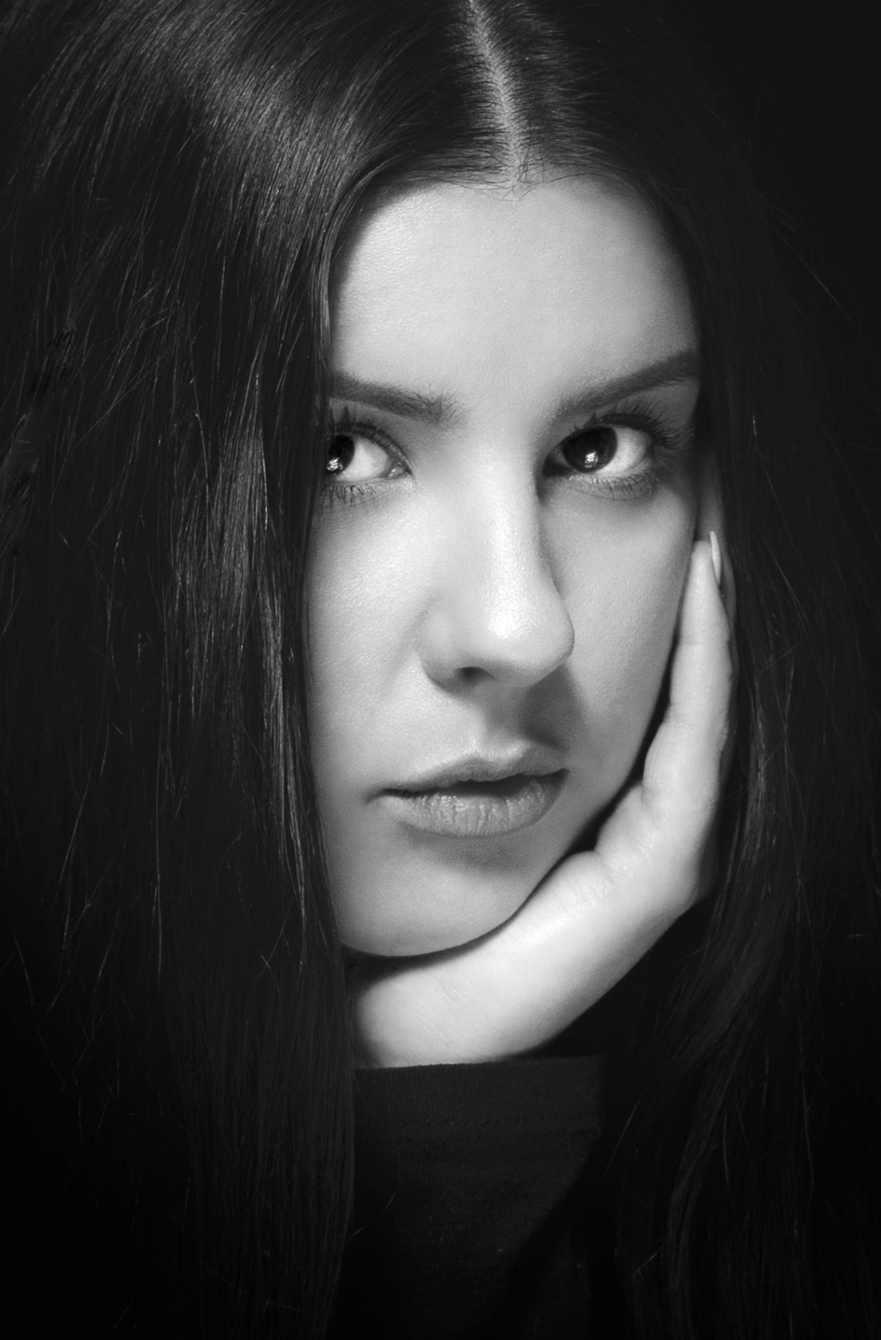 Czarno-biały portret kobiety z długimi, ciemnymi włosami, opierającej brodę na dłoni. Spojrzenie skierowane wprost, miękkie światło podkreśla rysy twarzy.