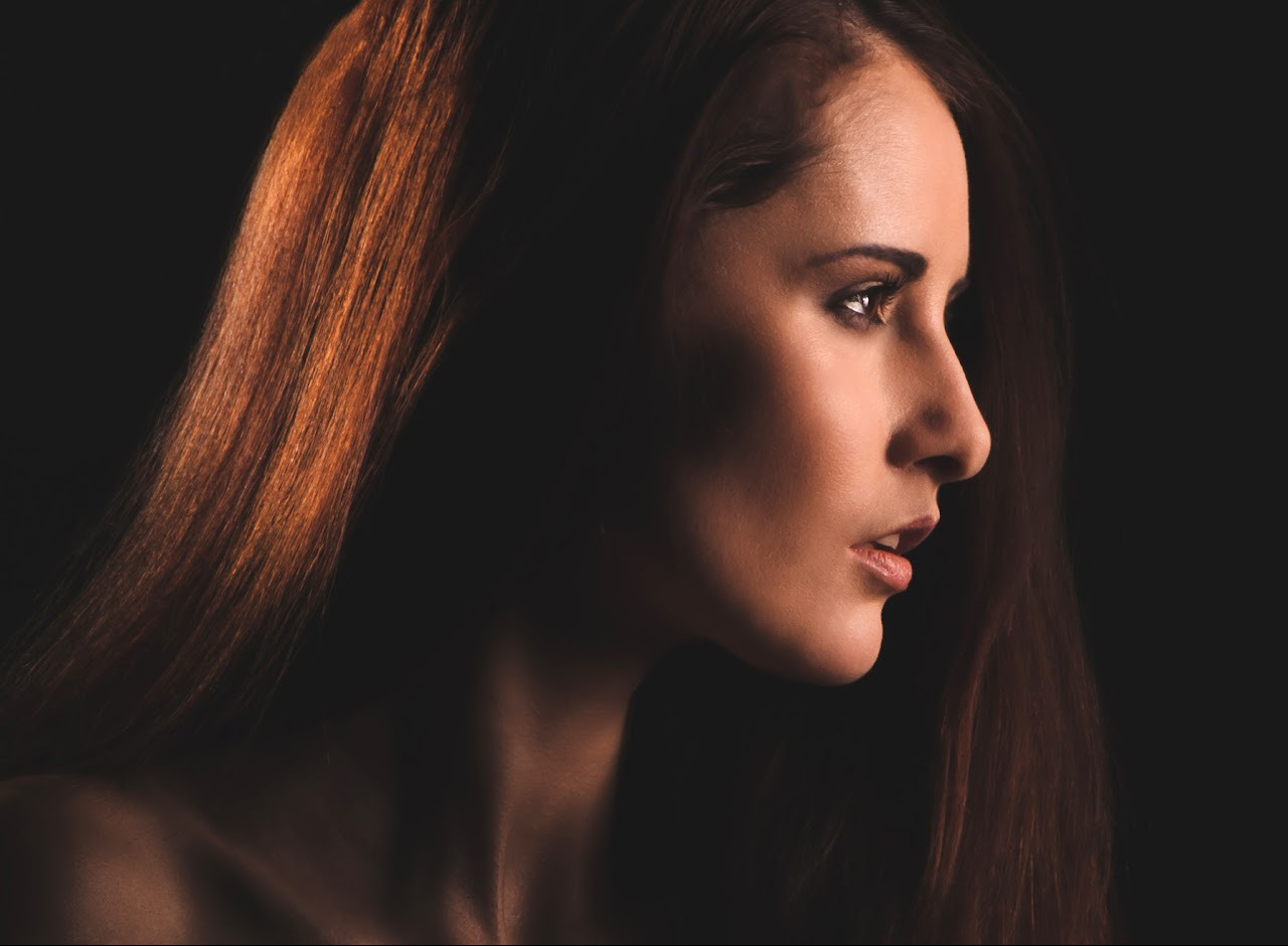 Portret kobiety z profilu, z długimi, rudymi włosami, oświetlonymi z góry, na ciemnym tle. Subtelny makijaż podkreśla rysy twarzy.