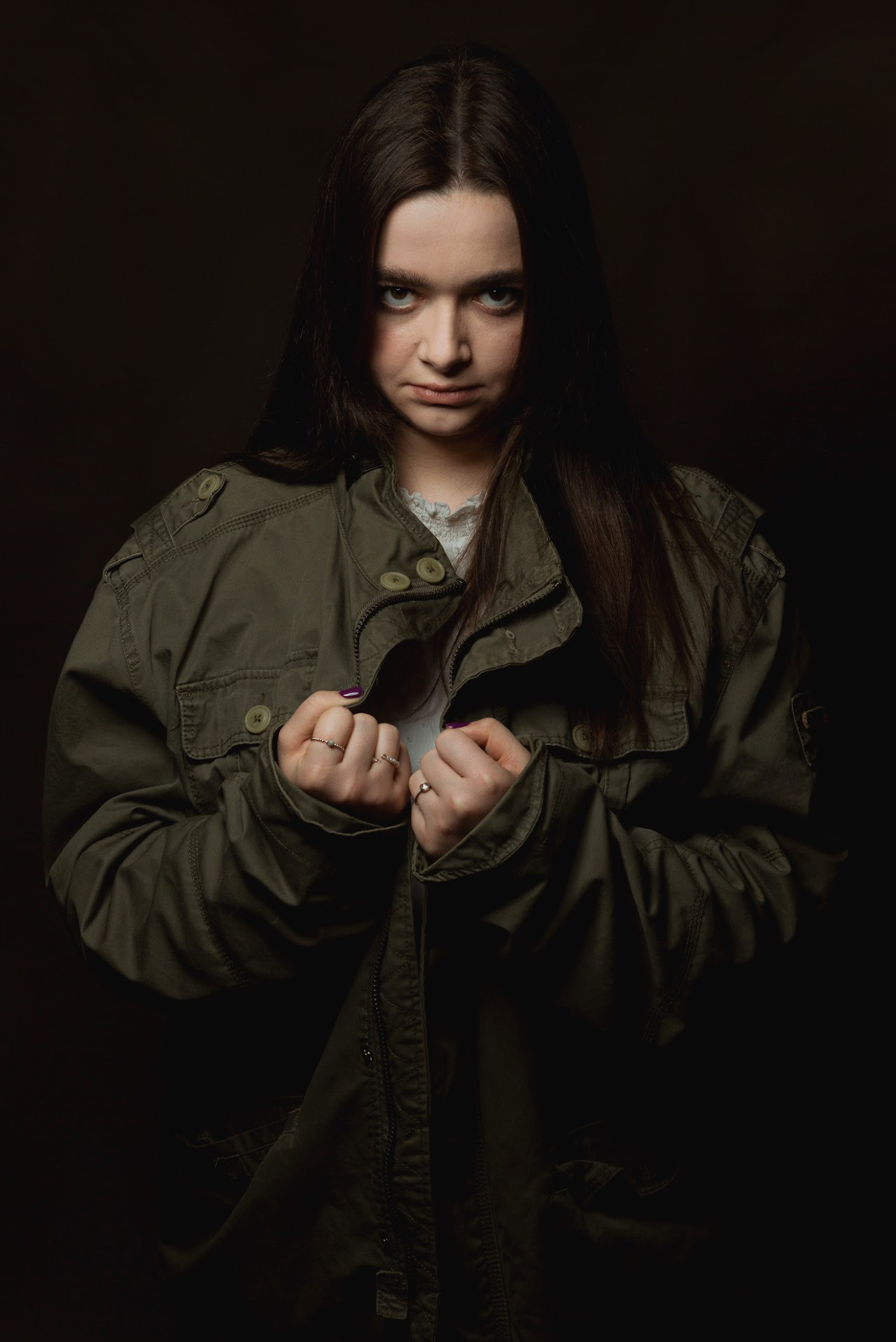 Portret młodej kobiety w zielonej kurtce na ciemnym tle. Uwagę przykuwają jej przenikliwe spojrzenie i dłonie z pierścionkami, trzymające poły kurtki.