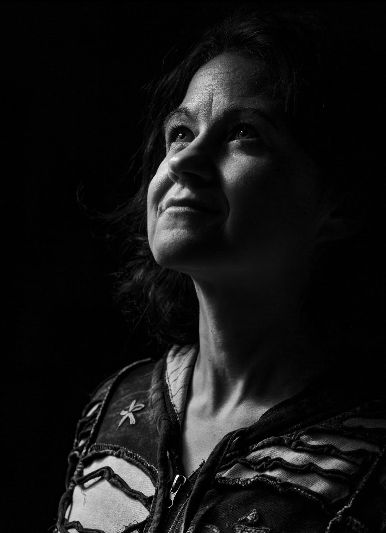 Czarno-biały portret kobiety spoglądającej w górę, z delikatnym uśmiechem i ciemnym tłem, podkreślającym kontury twarzy i fakturę ubrania.