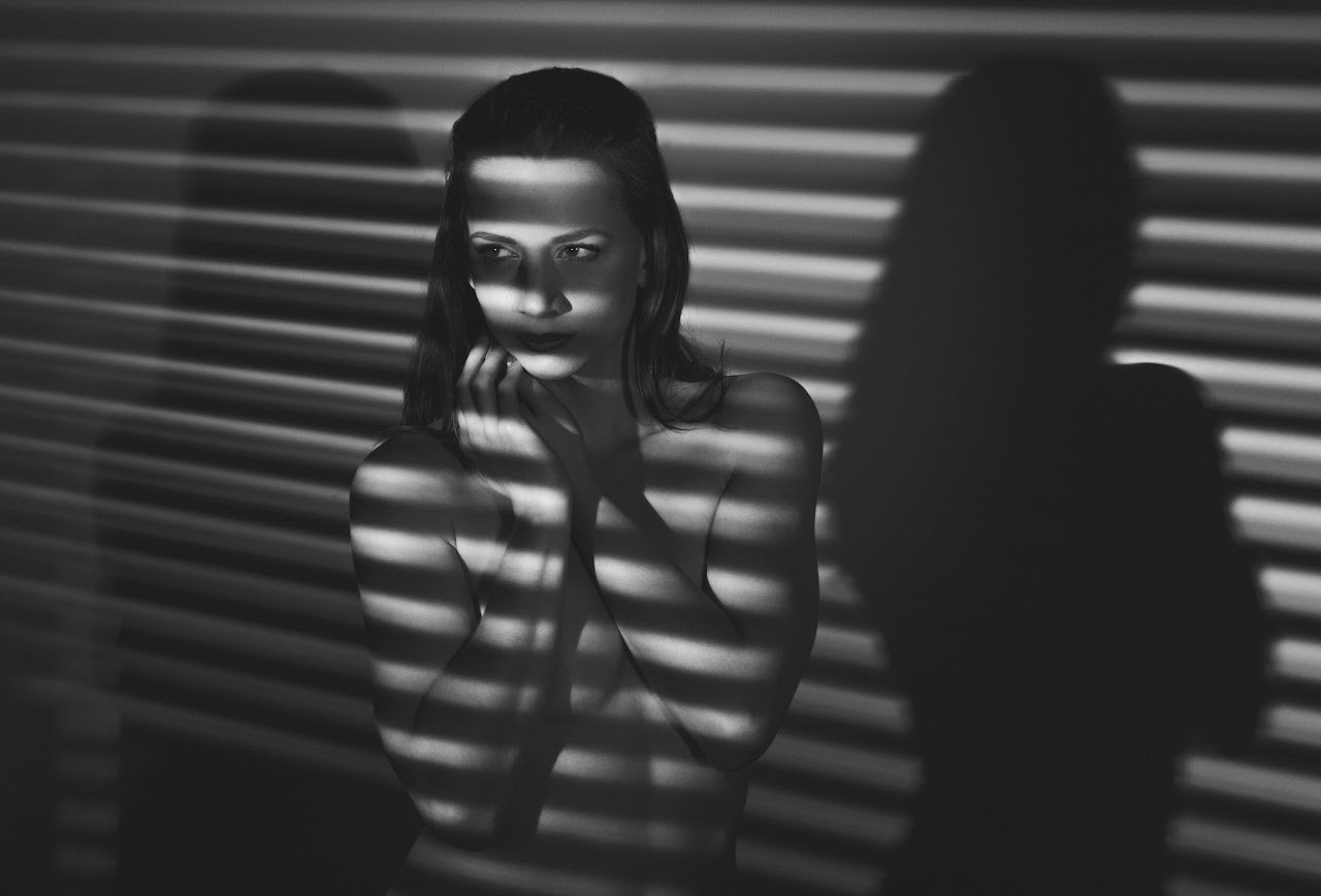 Czarno-biały portret kobiety w artystycznym ujęciu, z cieniem żaluzji tworzącym unikalny wzór na jej ciele i twarzy. Delikatny makijaż, zamyślony wyraz twarzy.