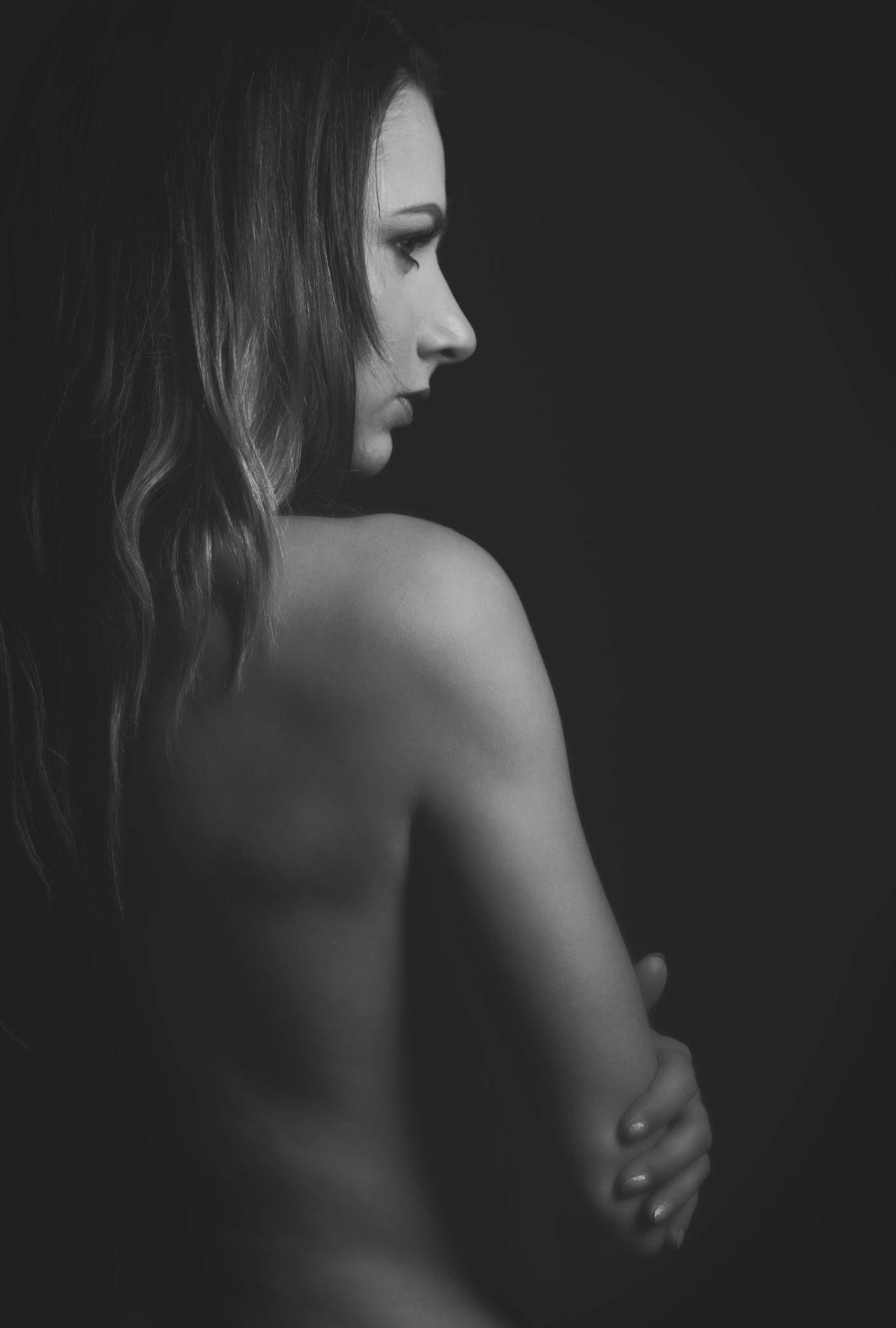 Czarno-biały portret kobiety z profilu, z odsłoniętymi plecami i ramionami, na ciemnym tle. Delikatne światło podkreśla kontury twarzy i sylwetki.