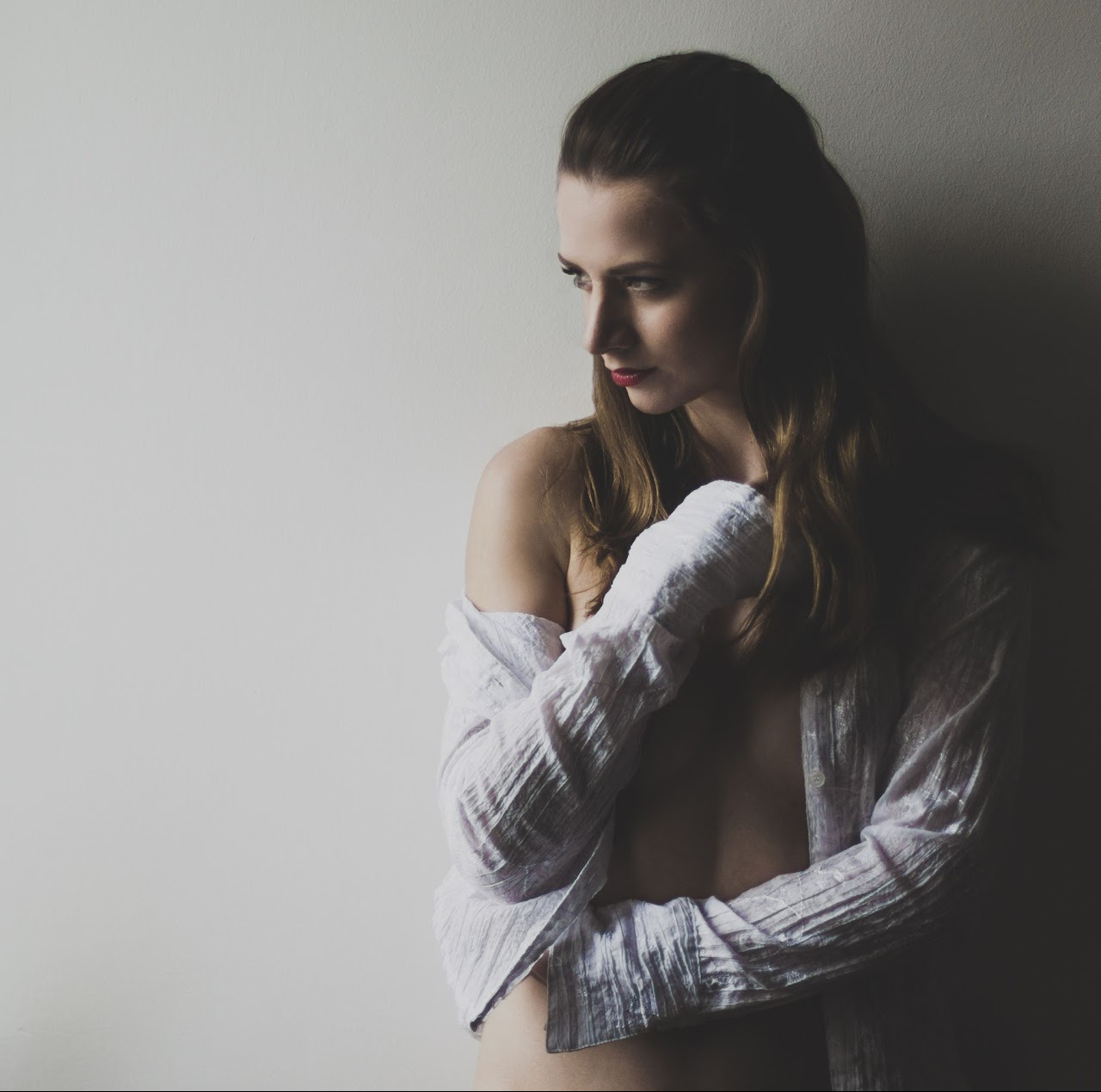 Portret kobiety w białej, rozpiętej koszuli, z delikatnym makijażem i spojrzeniem w bok, na tle jasnej ściany. Subtelne oświetlenie podkreśla rysy twarzy.