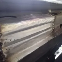 Naprawiona podłoga samochodu po spawaniu, widoczne nowe elementy blacharskie i spawy. Detal podwozia z bliska, zarys opony w tle.