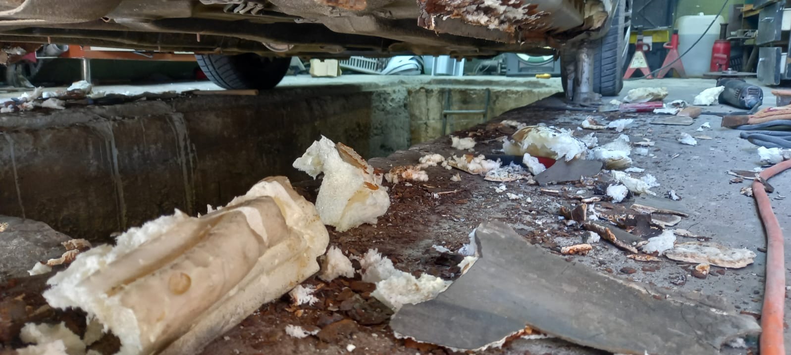 Uszkodzone podwozie samochodu na podnośniku w warsztacie, widoczne fragmenty skorodowanej blachy i pianki montażowej. Niefachowa naprawa antykorozyjna.
