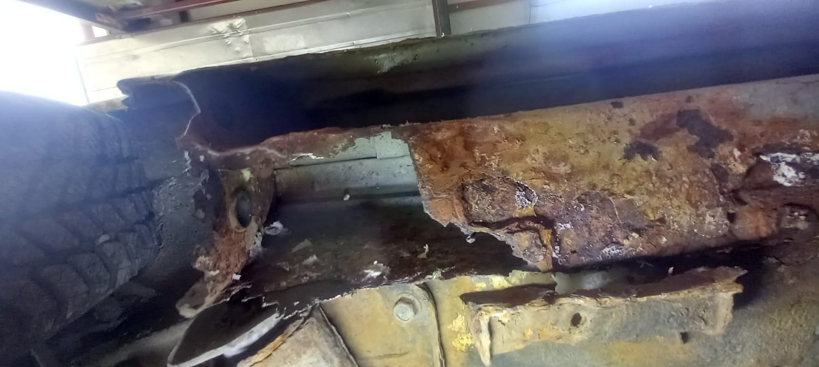 Zniszczony, skorodowany próg samochodu z widocznymi śladami rdzy i ubytkami metalu. Fragment podwozia z elementami mocującymi. Niejednolite oświetlenie.