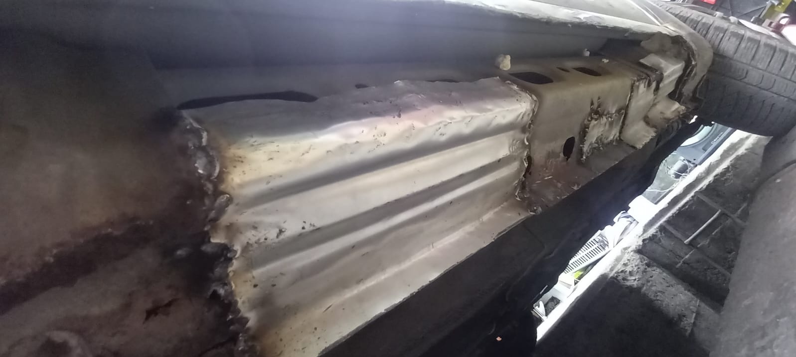 Naprawiona podłoga samochodu po spawaniu, widoczne nowe elementy blacharskie i spawy. Detal podwozia z bliska, zarys opony w tle.