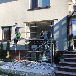 Home & Trade Tomasz Gliński - Demontaż okna na rusztowaniu. Dwóch pracowników w trakcie prac przygotowawczych do montażu nowego okna. Gruz i resztki materiałów budowlanych na ziemi.