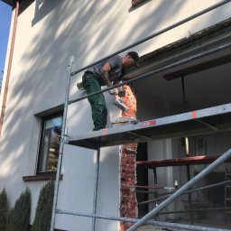 Home & Trade Tomasz Gliński - Pracownik na rusztowaniu, w trakcie skuwania tynku z elewacji domu jednorodzinnego. Widoczne narzędzia i fragment odsłoniętej cegły. Słoneczny dzień.