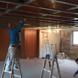 Home & Trade Tomasz Gliński - Pracownik na drabinie montuje metalowy stelaż sufitu podwieszanego w remontowanym pomieszczeniu. Widoczne elementy instalacji i surowy stan ścian.